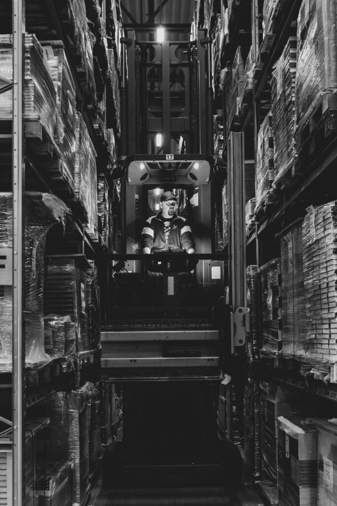A worker operates a forklift in a warehouse, surrounded by tall shelves of packaged goods.